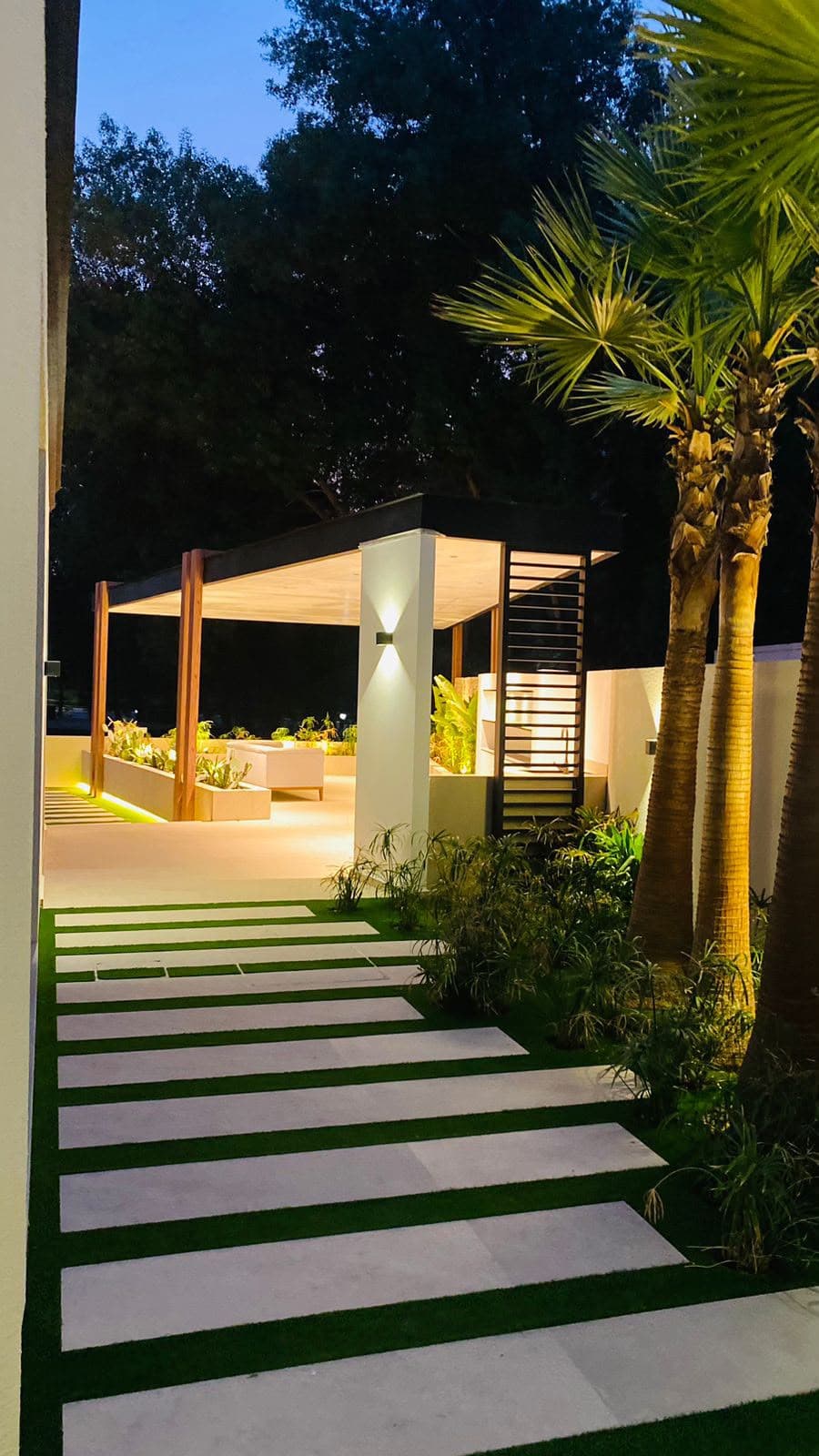 Stone pathway leading to a lit aluminium pergola through palm trees